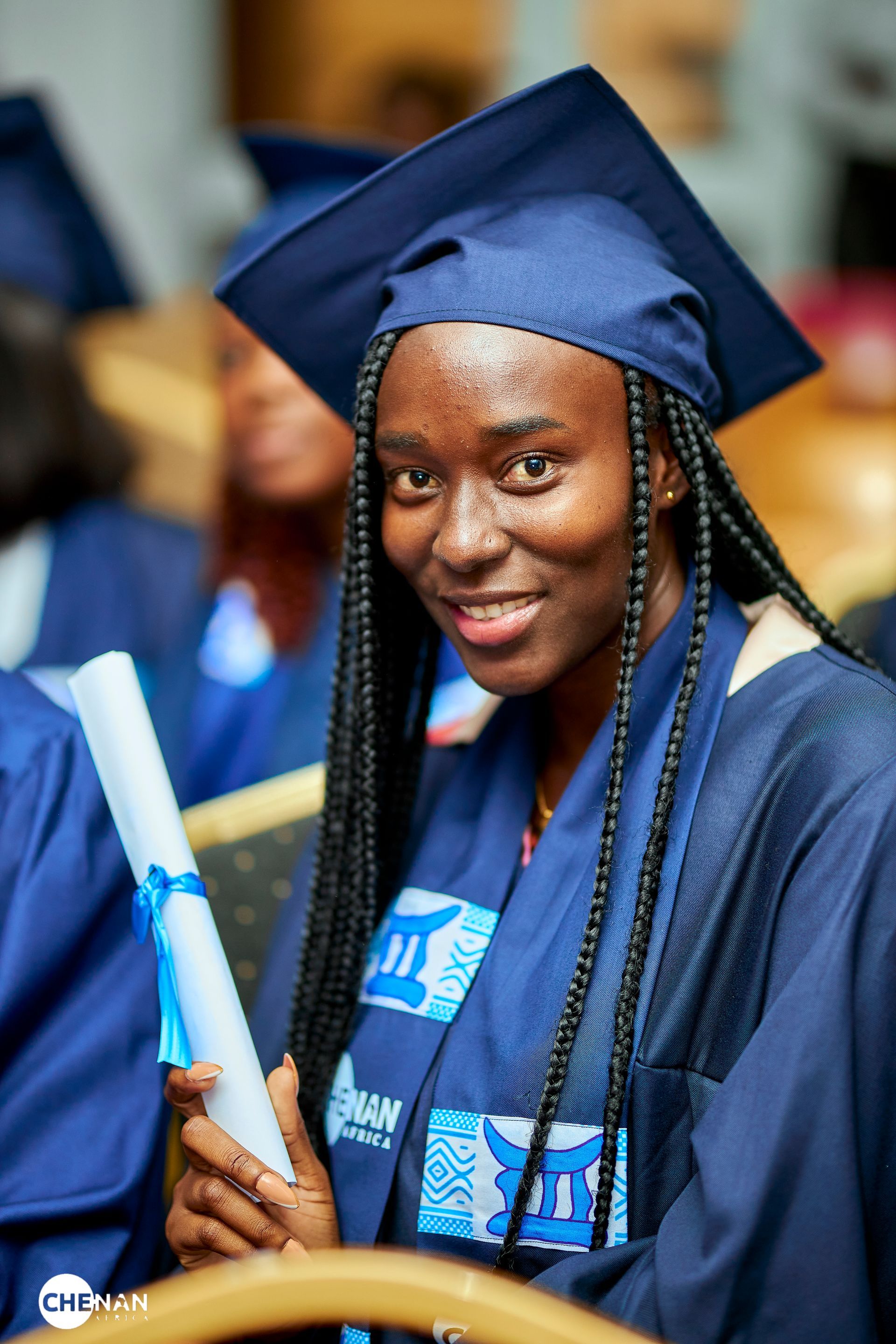 Une étudiante diplômée, coiffée d'une toque bleue et vêtue d'une robe de cérémonie, sourit en tenant son diplôme, à l'intérieur.