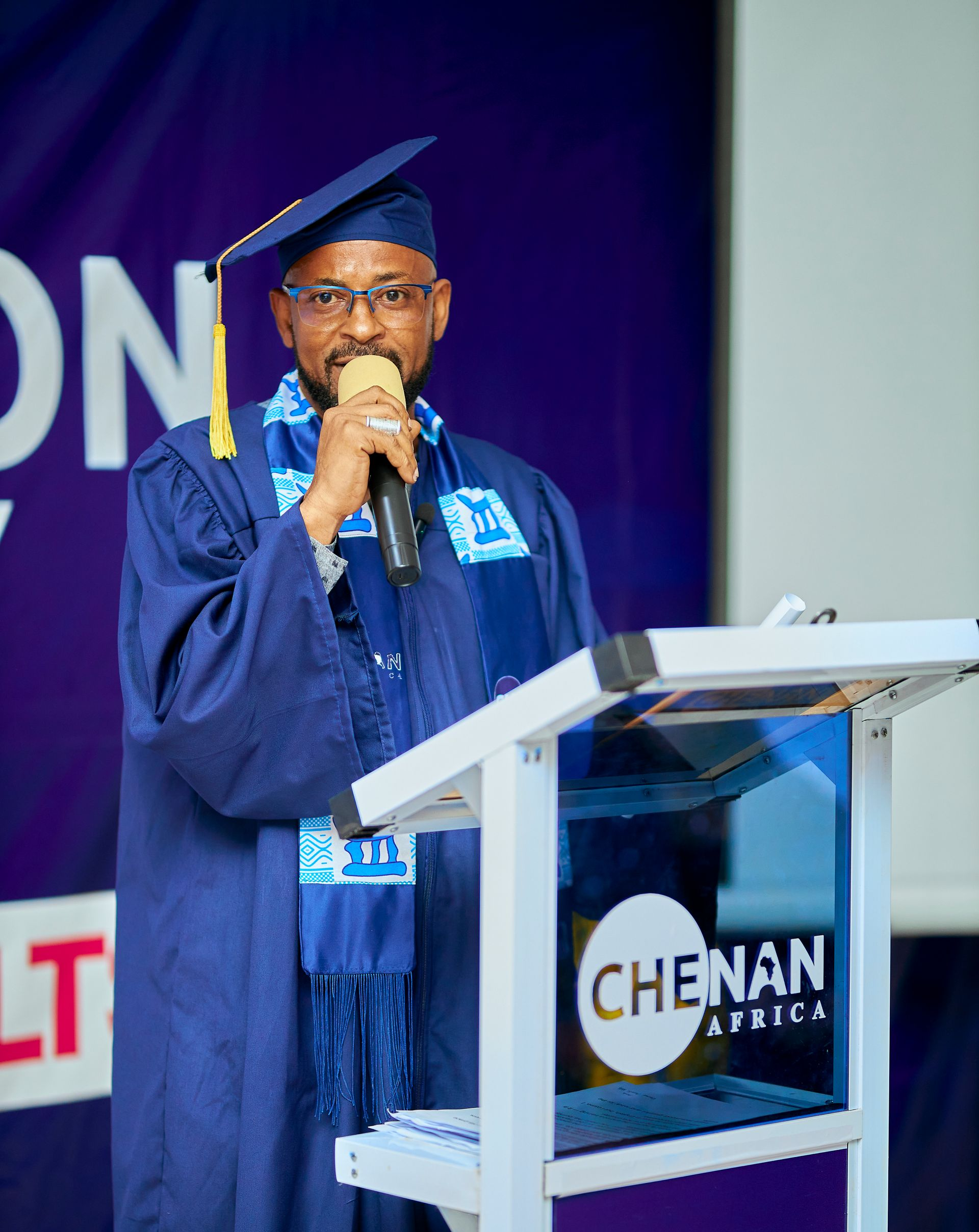 Un homme en toge bleue de remise de diplôme prend la parole à un podium orné du logo « CHENAN AFRICA ».