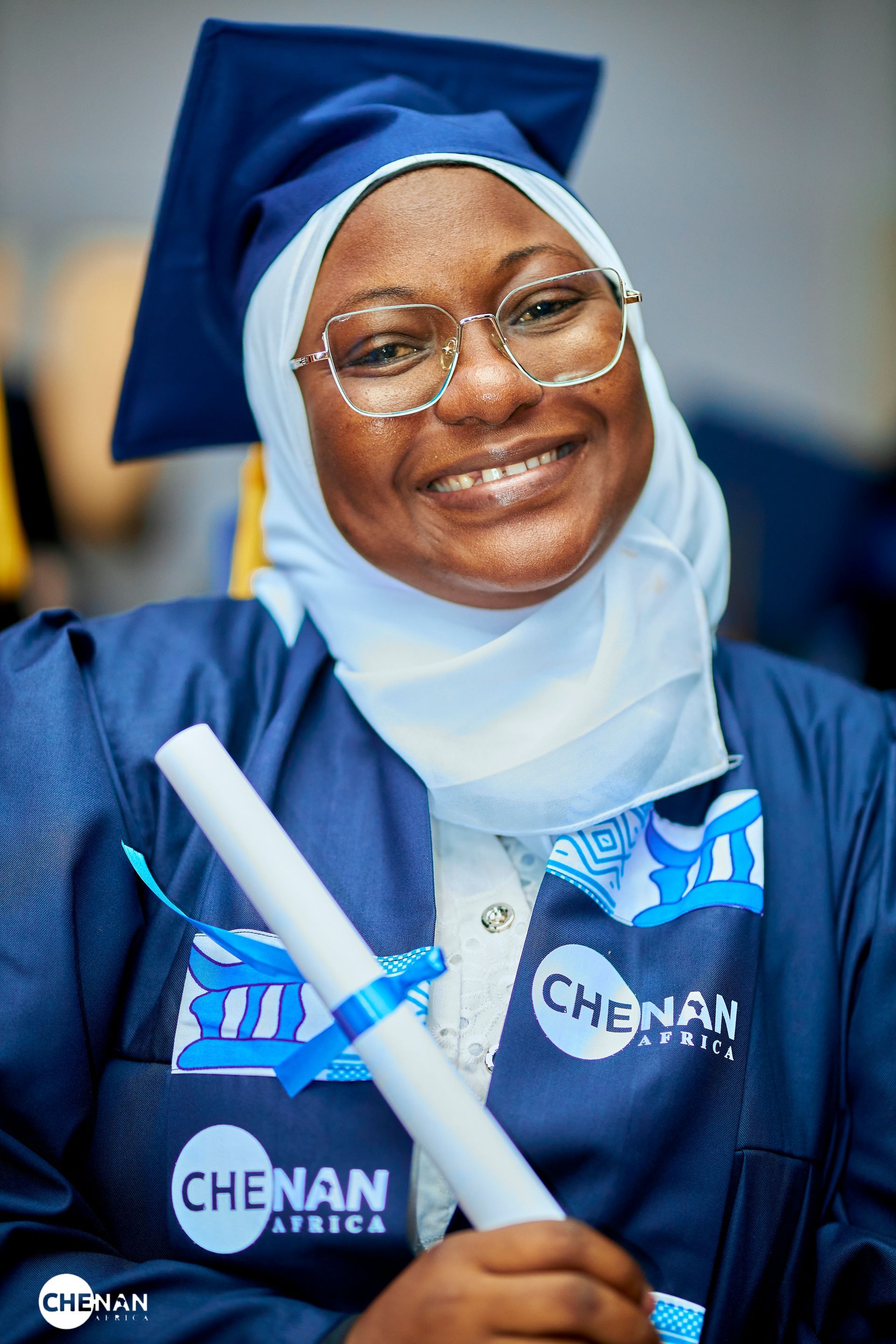 Une femme en tenue de remise de diplôme, tenant son diplôme et souriante, porte un hijab, des lunettes, une toque et une robe bleues.
