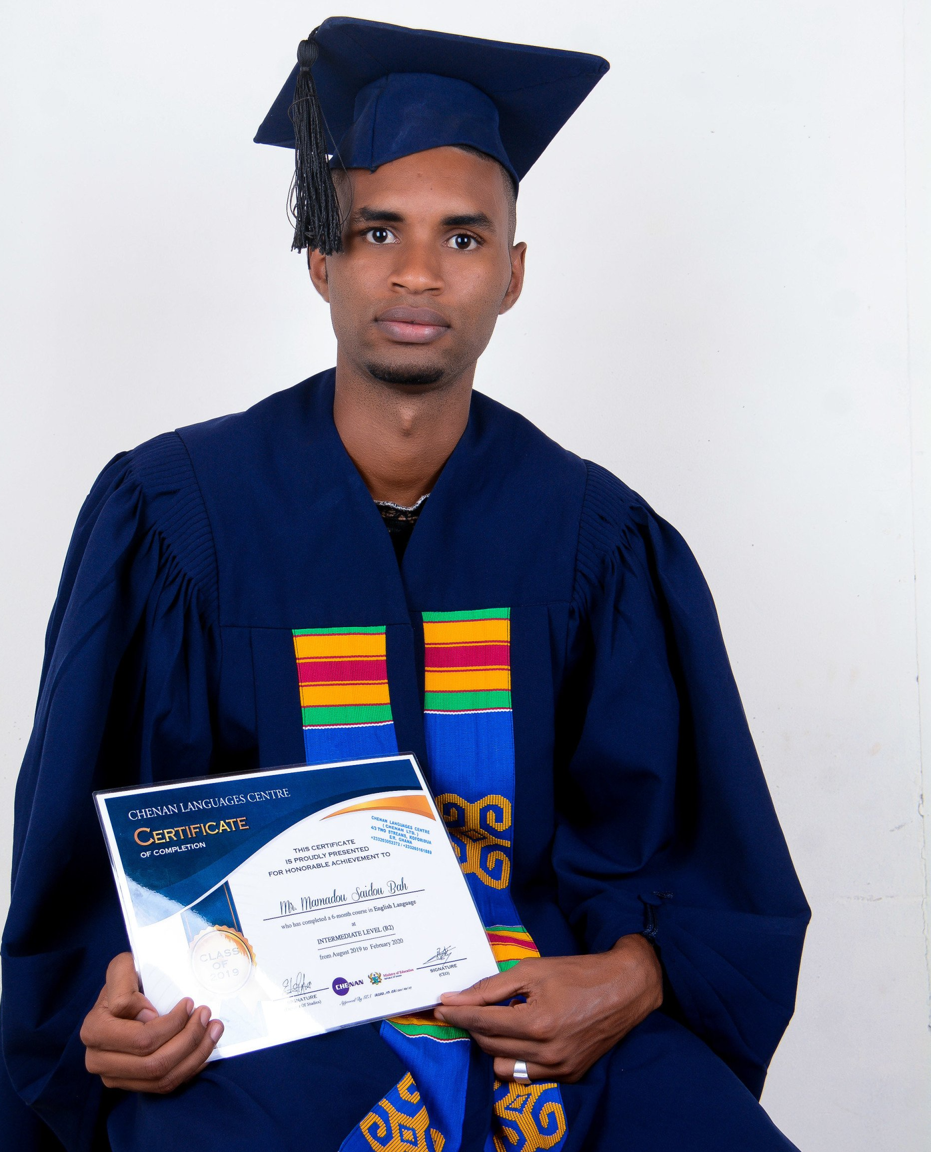 Un homme en tenue de remise de diplôme tient son diplôme ; toque bleue, toque, écharpe rouge et or.