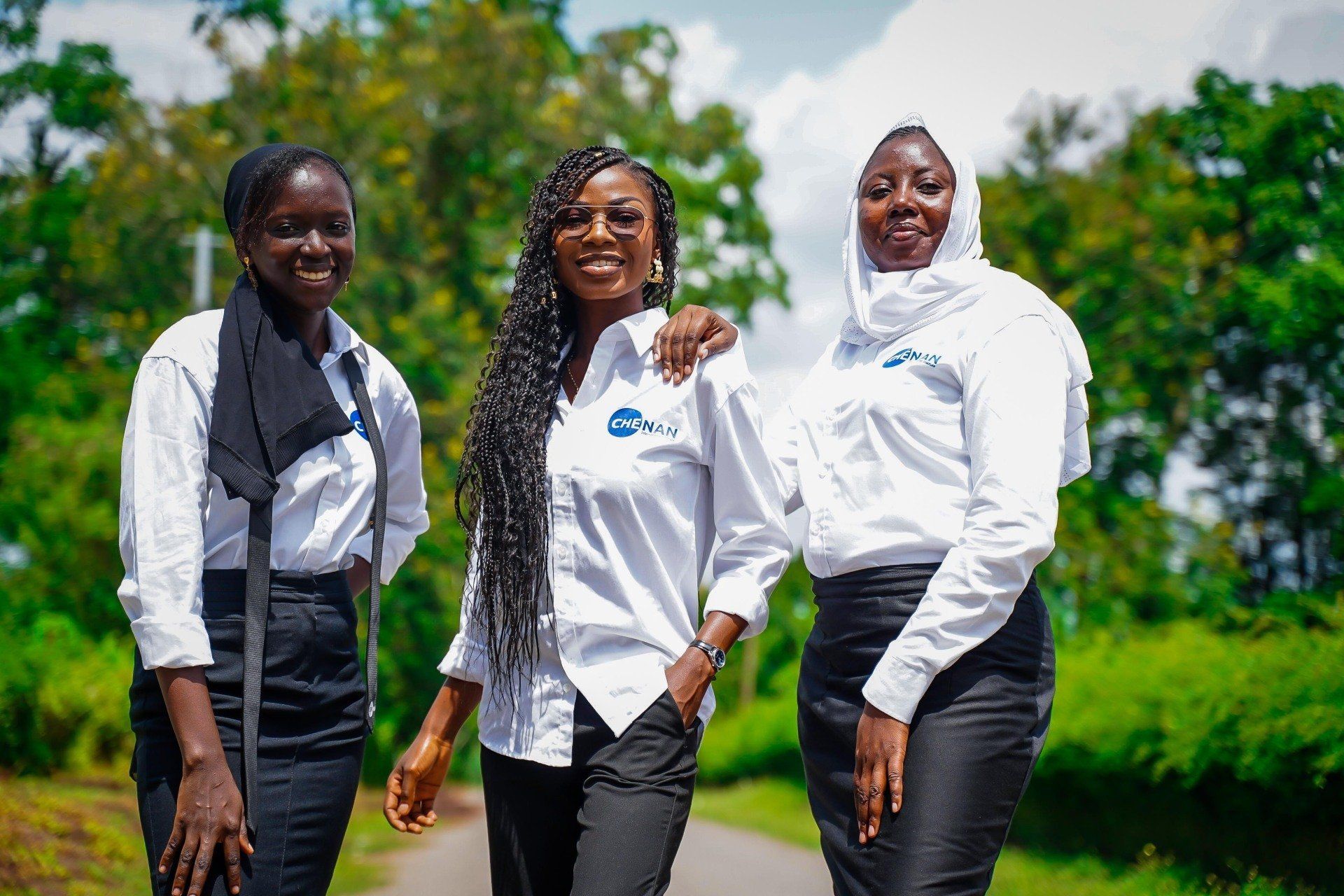 Trois femmes se tiennent au bord d'une route, souriantes. Elles portent des chemises blanches à boutons avec un logo et des jupes noires, dans un décor extérieur verdoyant.
