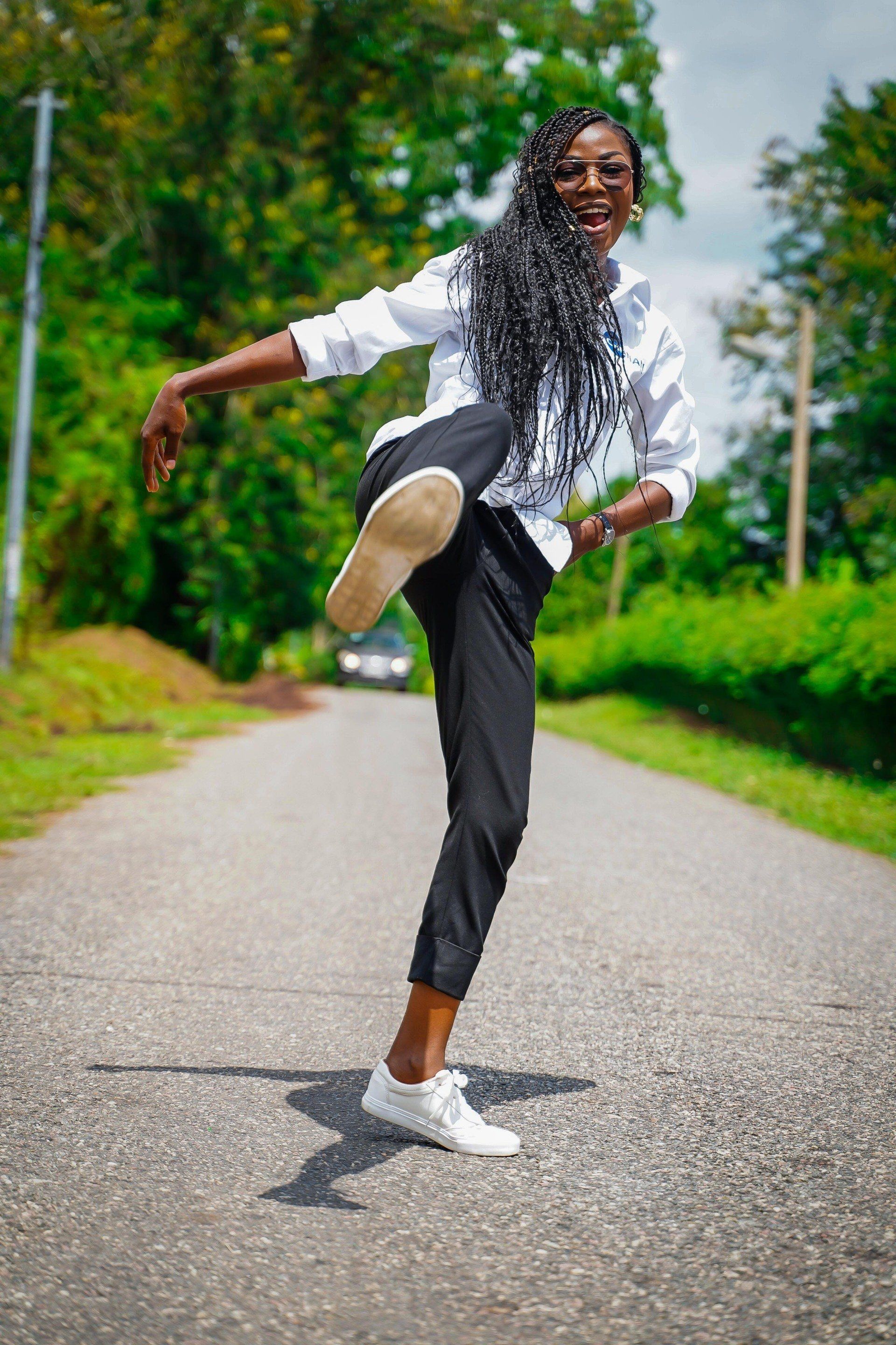 Une femme levant la jambe sur la route, vêtue d'une chemise blanche, d'un pantalon noir, de baskets blanches et de longs cheveux tressés.