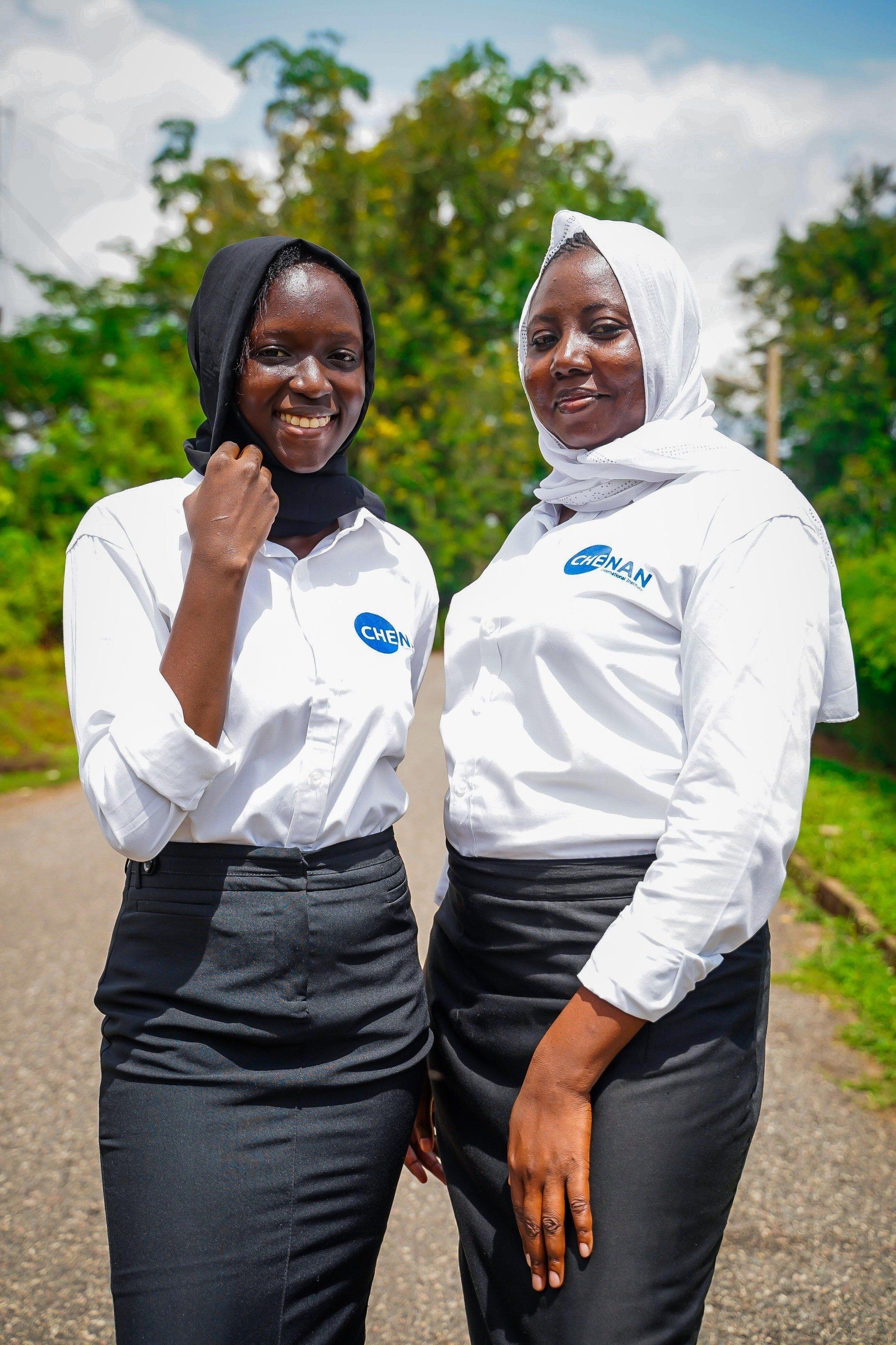 Deux femmes en chemises blanches et jupes noires, debout à l'extérieur, souriantes, l'une d'elles ajustant son hijab.