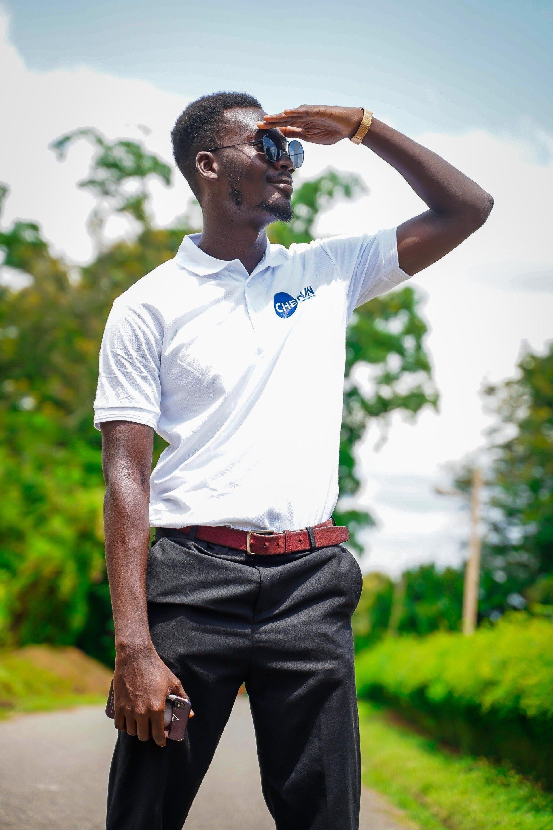 Un homme en chemise blanche et lunettes de soleil regarde droit devant lui, les yeux protégés. Il se tient sur le bord de la route.