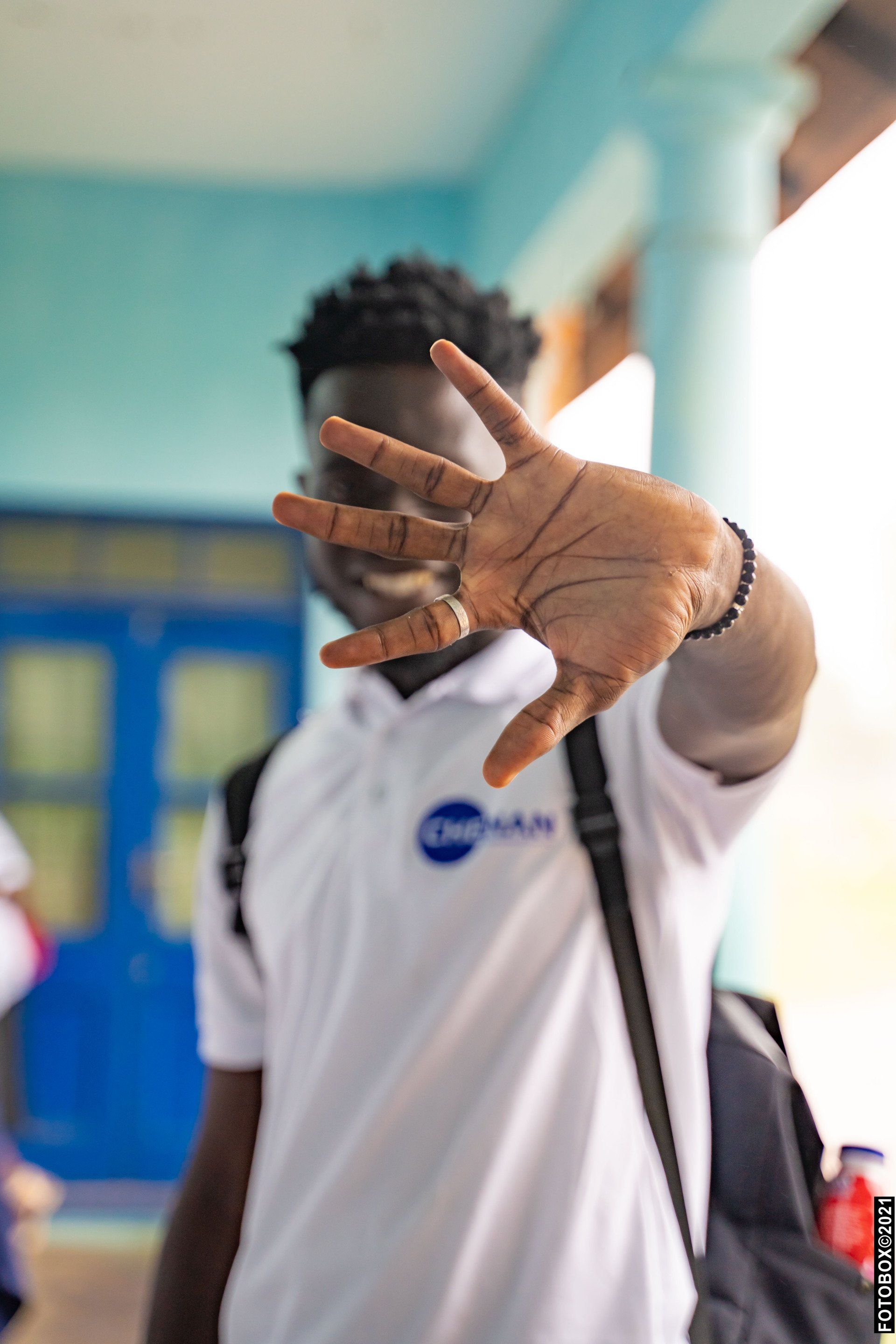 Une personne tend la main vers la caméra, obstruant la vue. Elle porte un t-shirt blanc avec un logo et un sac à dos.