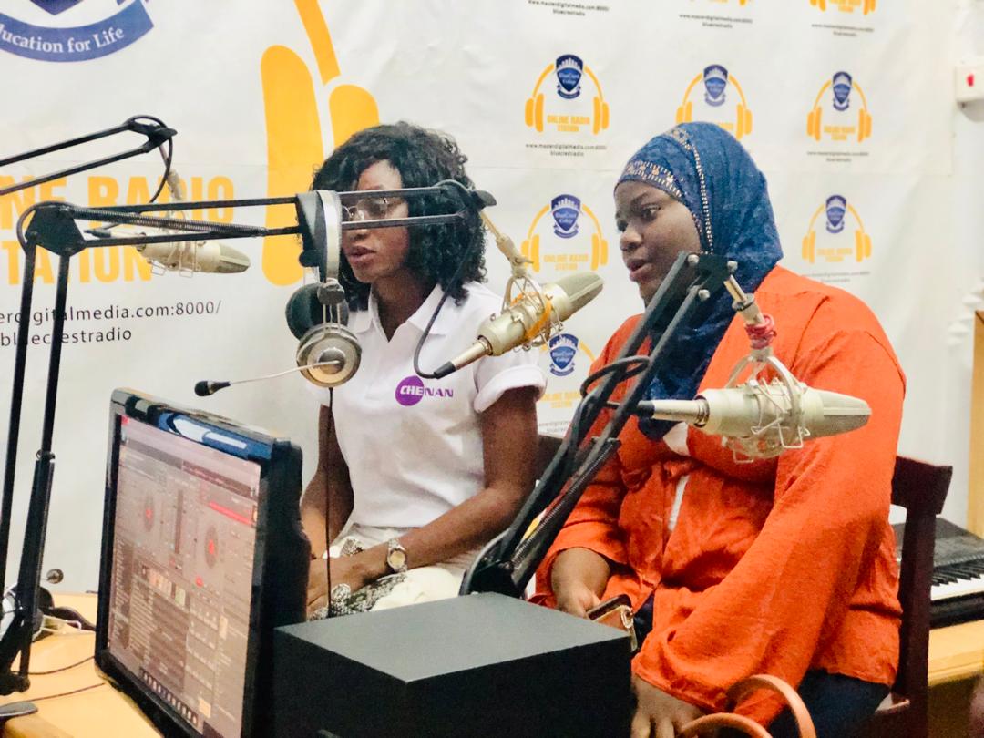 Deux femmes dans une station de radio ; l'une en chemise blanche et lunettes, l'autre portant un hijab, parlent dans des microphones.