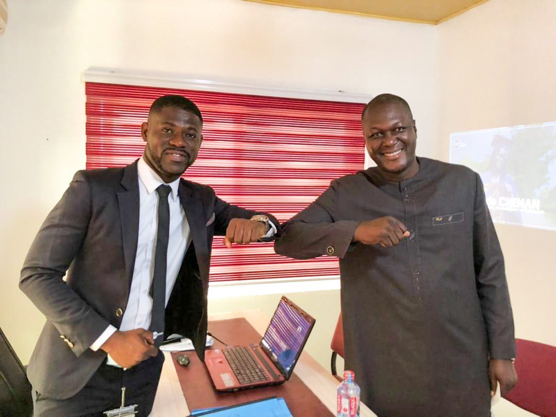 Deux hommes se saluent d'un coup de coude dans un bureau. L'un porte un costume, l'autre une tenue traditionnelle. Un ordinateur portable est posé sur le bureau, des stores rouges sont installés.