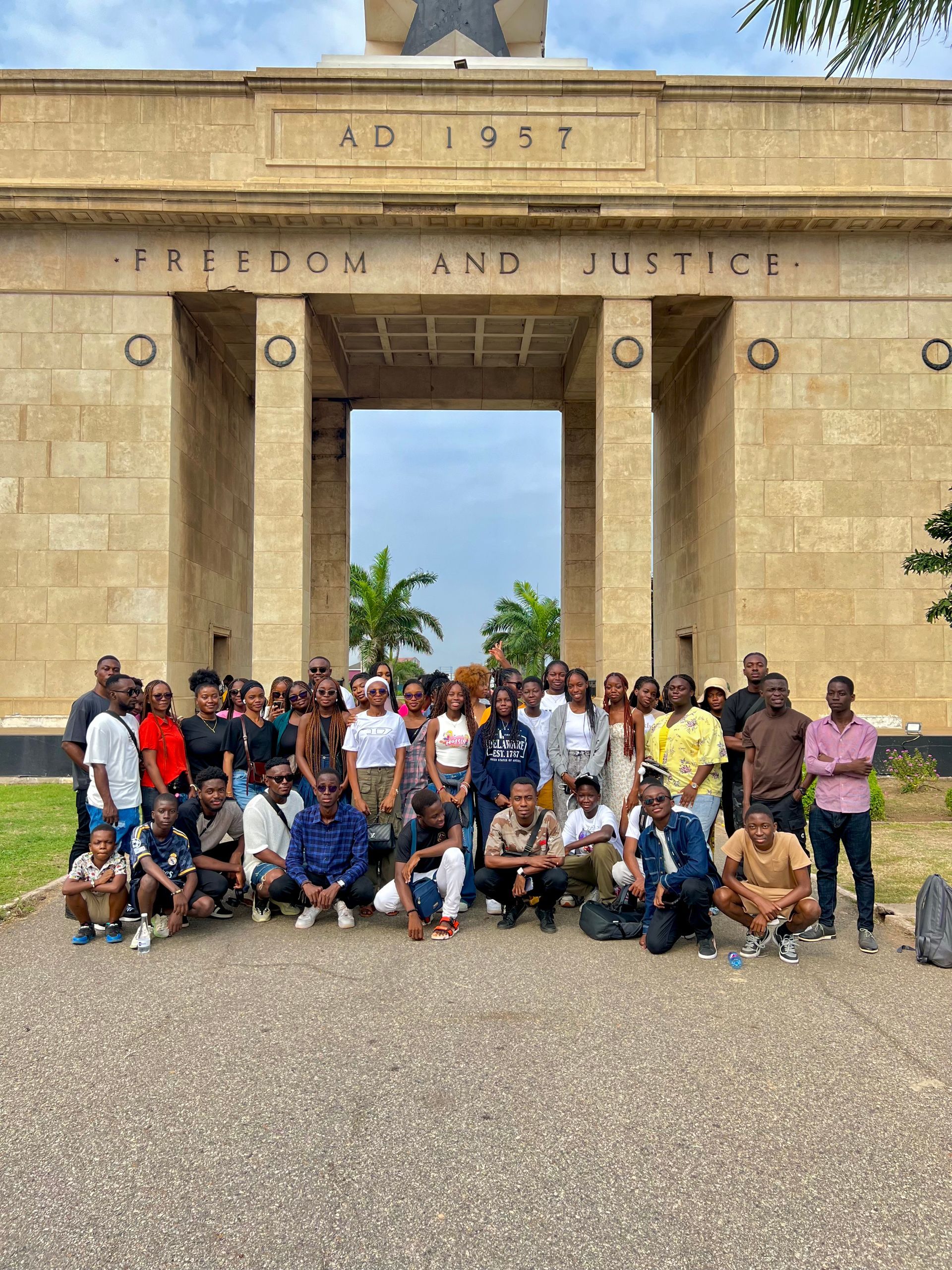 Un groupe important de personnes pose devant un monument où l'on peut lire « Liberté et Justice ». Ciel nuageux.