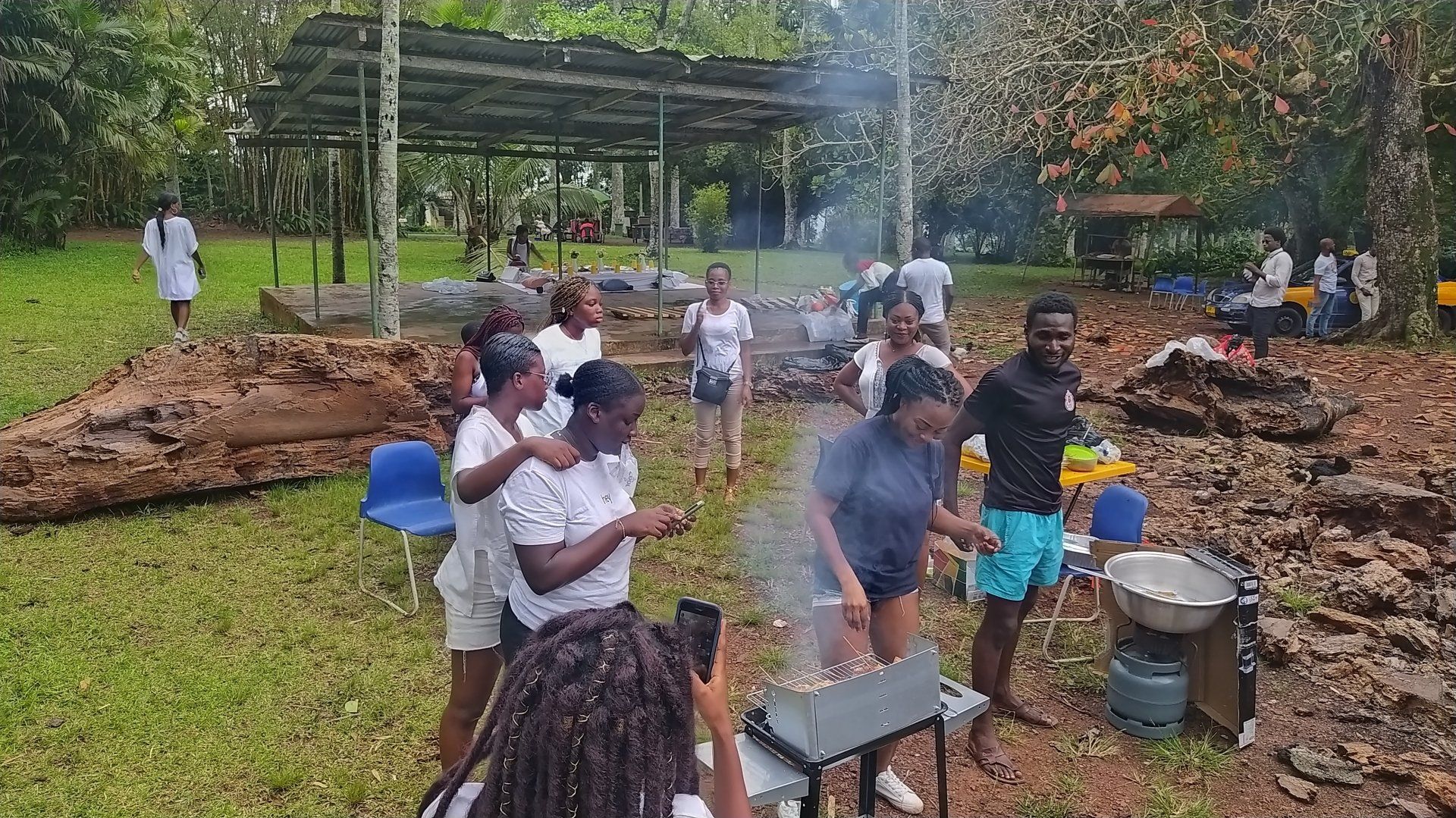 Un groupe de personnes rassemblées à l'extérieur préparent à manger sur un feu, sous un abri dans une zone boisée.