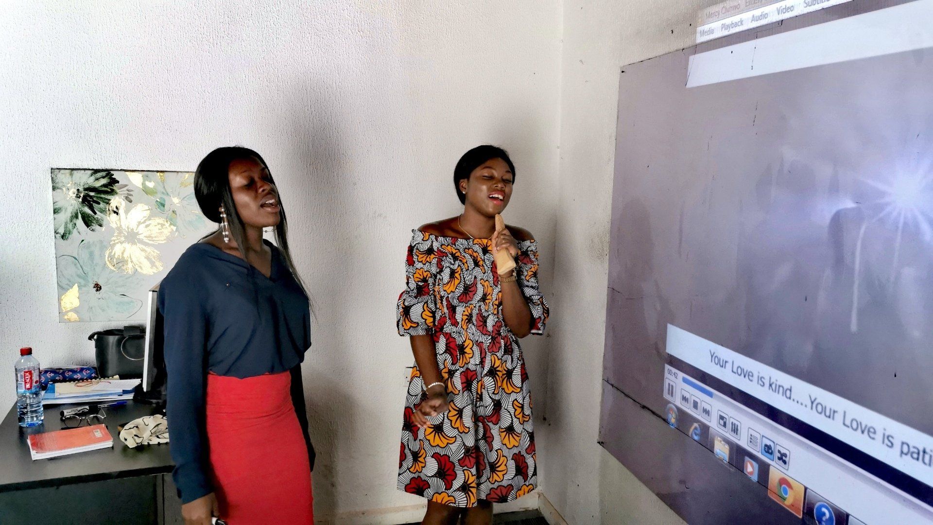Deux femmes chantent devant un écran affichant du texte et une lumière vive. L'une porte une jupe rouge et l'autre une robe à motifs.