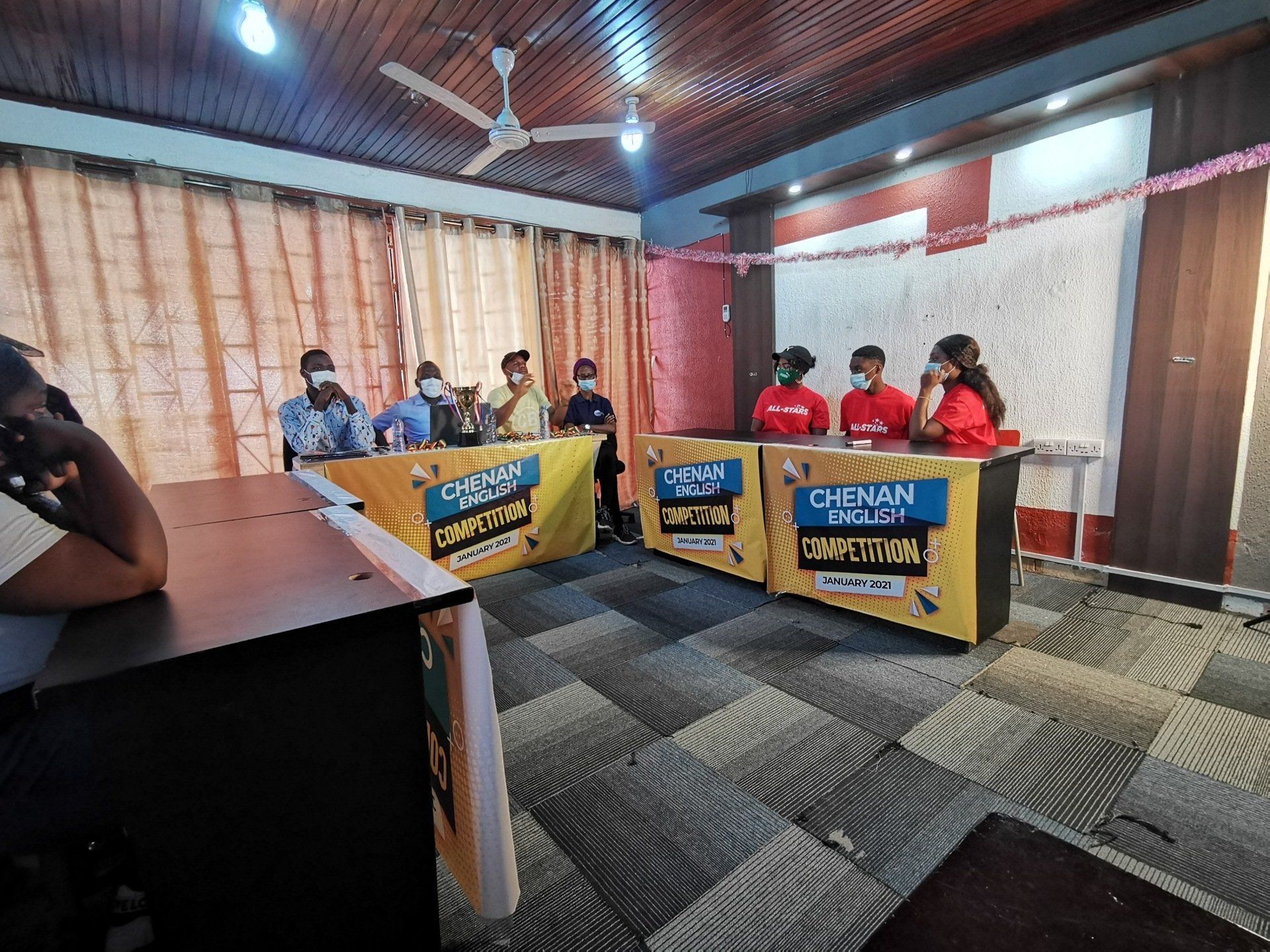 Des personnes sont assises à des tables dans une salle de réception. Certaines portent des masques. Des banderoles « Concours des écoles de Chennai » ornent les tables.
