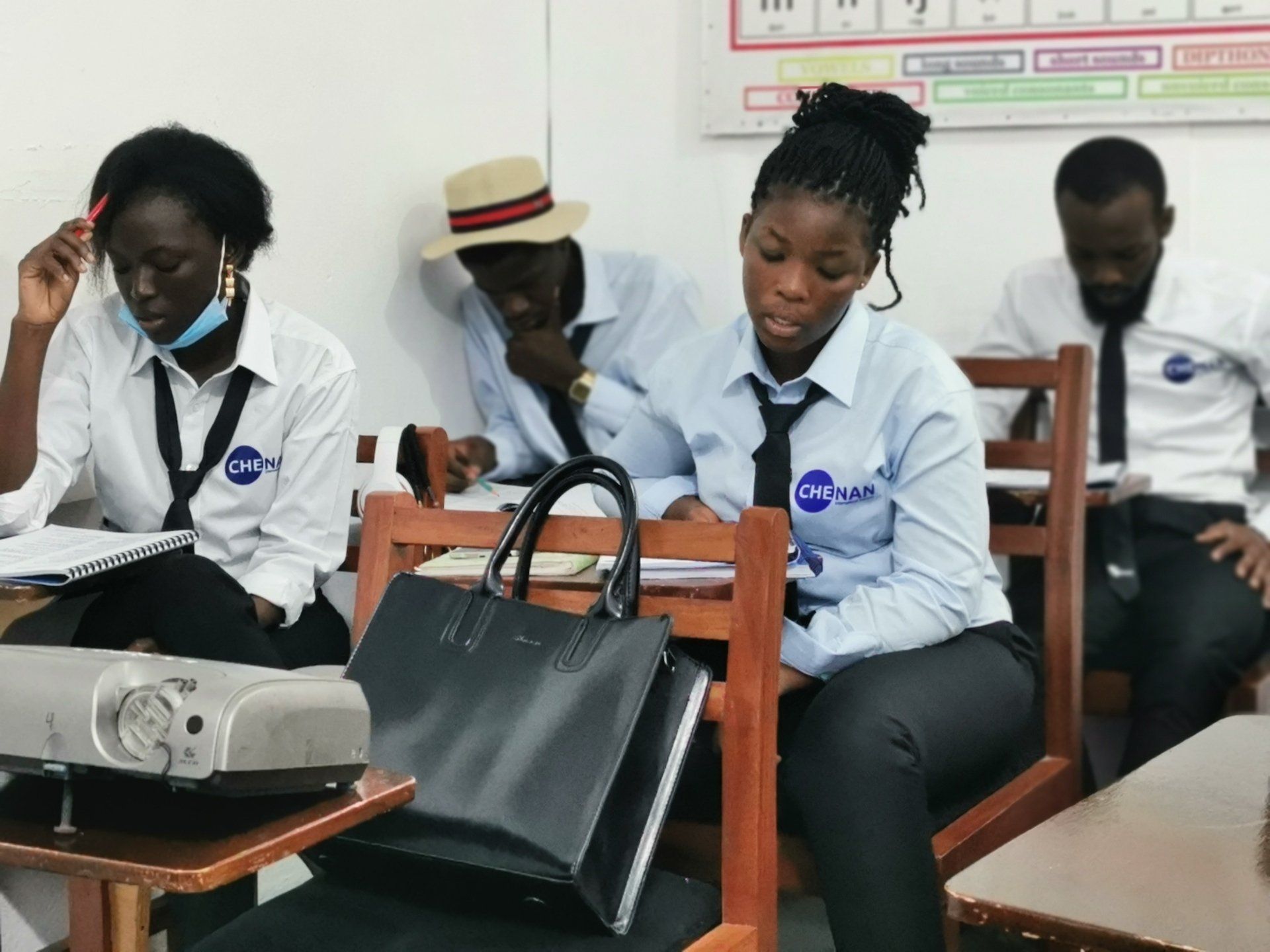 Dans une salle de classe, des élèves, vêtus de chemises d'uniforme bleu clair et de cravates, consultent des documents ou un ordinateur portable. Un projecteur est posé sur un bureau.