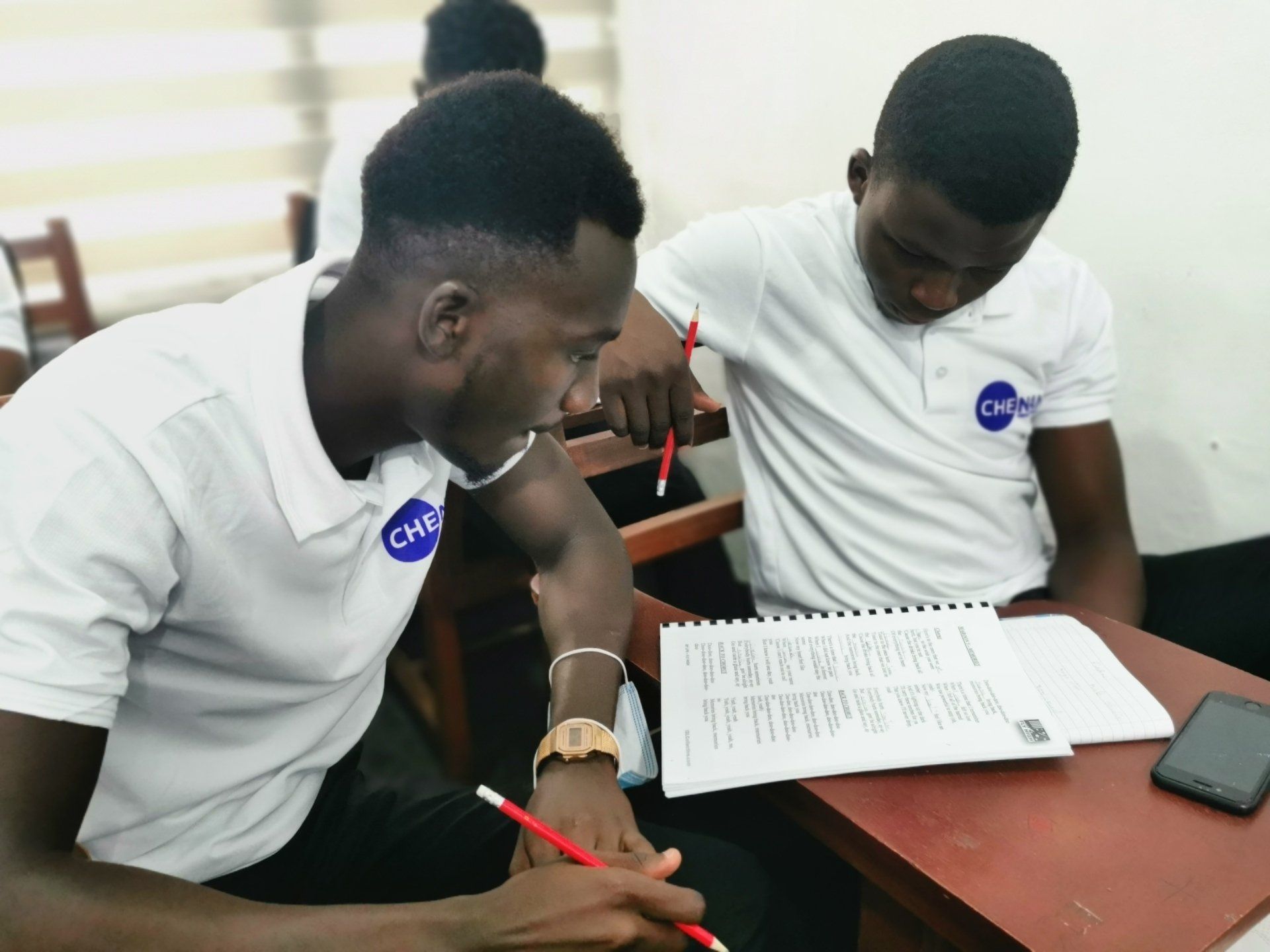 Deux jeunes hommes en chemise blanche étudient ensemble à un bureau, les yeux rivés sur un cahier et un smartphone.