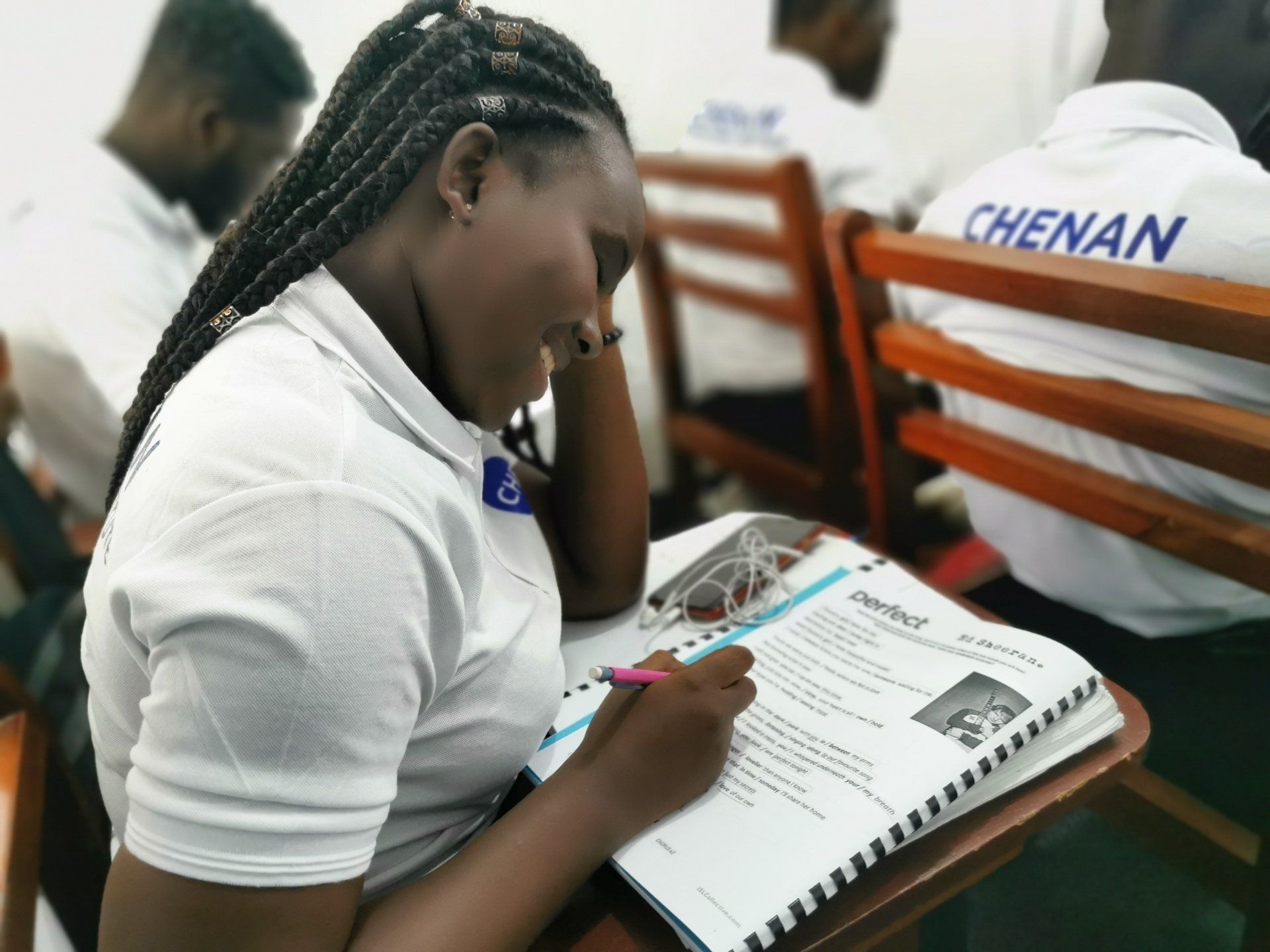 Personne en chemise blanche, tresses, écrivant dans un cahier à son bureau en classe.