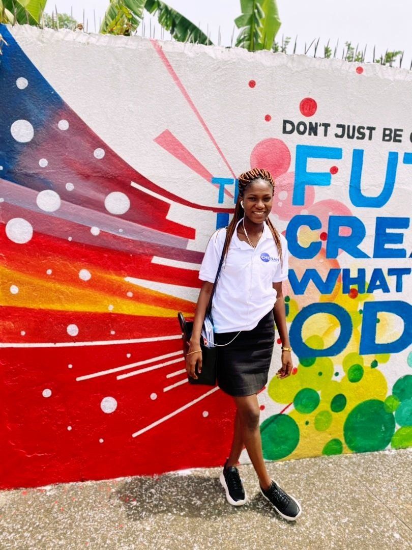 Une femme se tient devant une fresque colorée où l'on peut lire : « CRÉEZ VOTRE AVENIR ». Elle porte une chemise blanche, une jupe noire et des baskets.