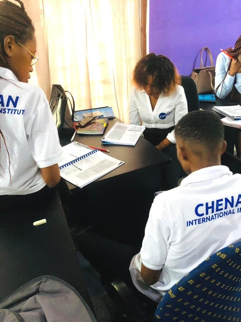 Des personnes vêtues de chemises blanches portant l'inscription « Chenan International Institute » étudient des documents à un bureau dans une salle de classe.
