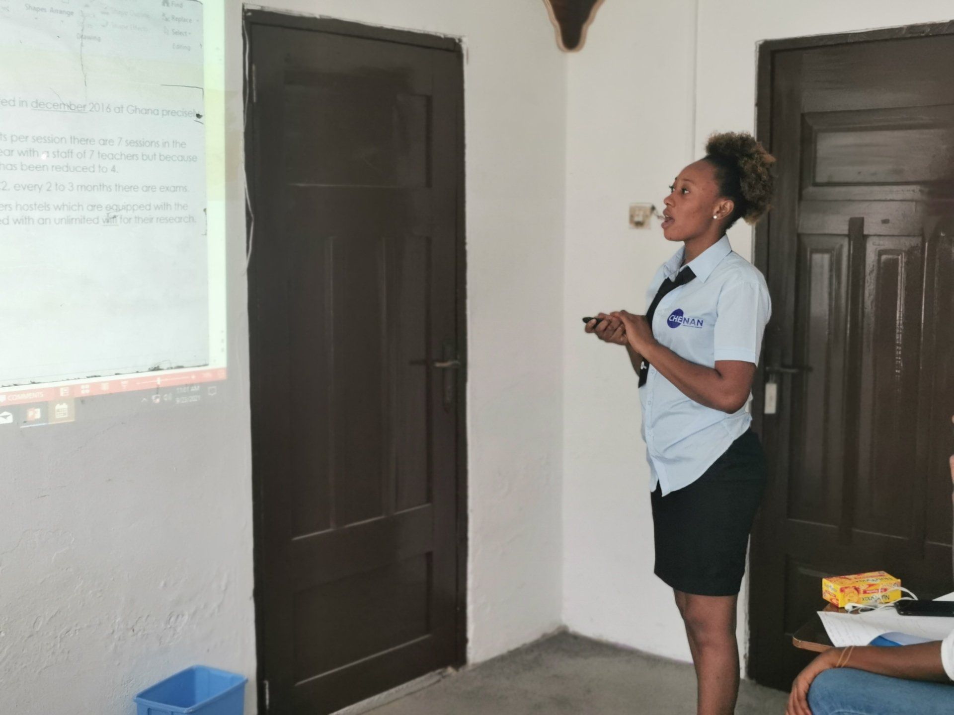 Femme faisant une présentation, tenant une télécommande, près d'un écran et d'une porte fermée.