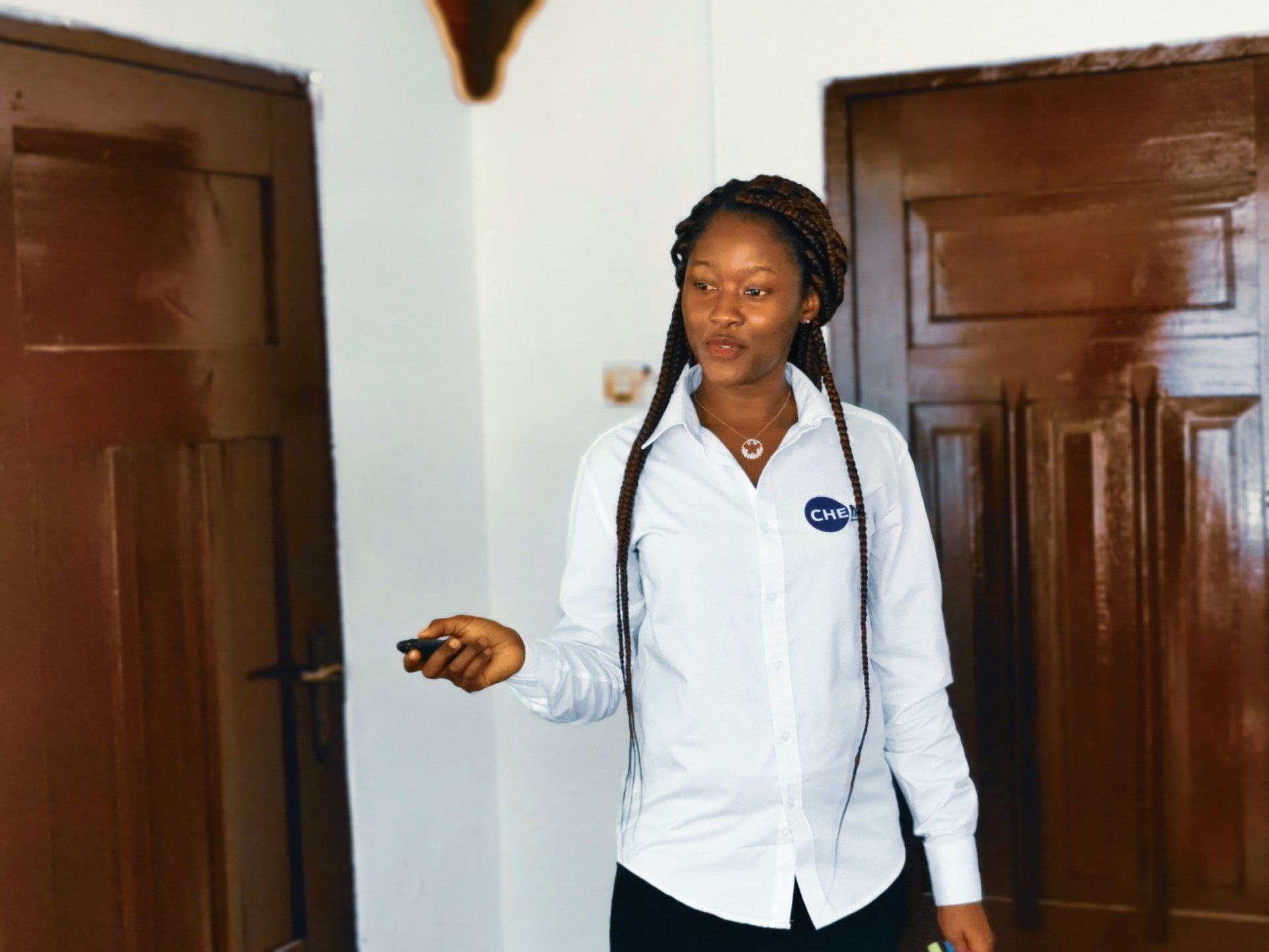 Une femme en chemise blanche tenant une télécommande se tient entre deux portes marron.
