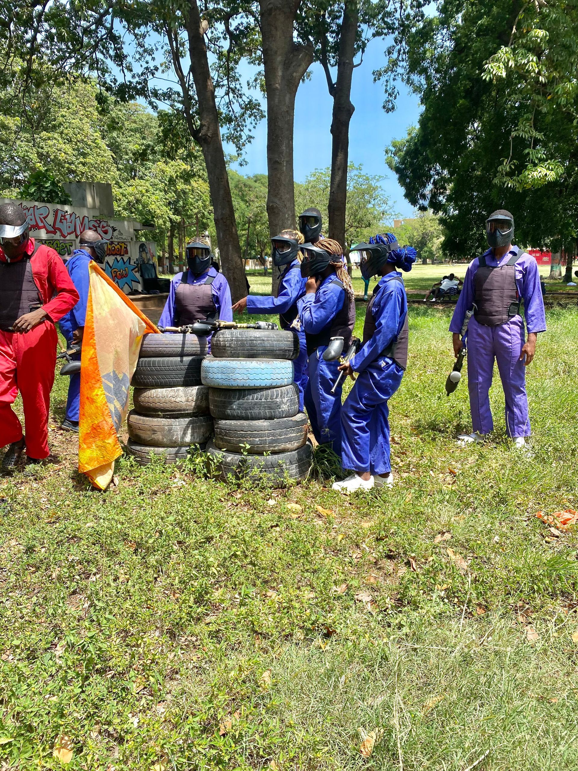 Des personnes en tenue de protection jouent au paintball dans une zone herbeuse parsemée d'obstacles de pneus et d'arbres.