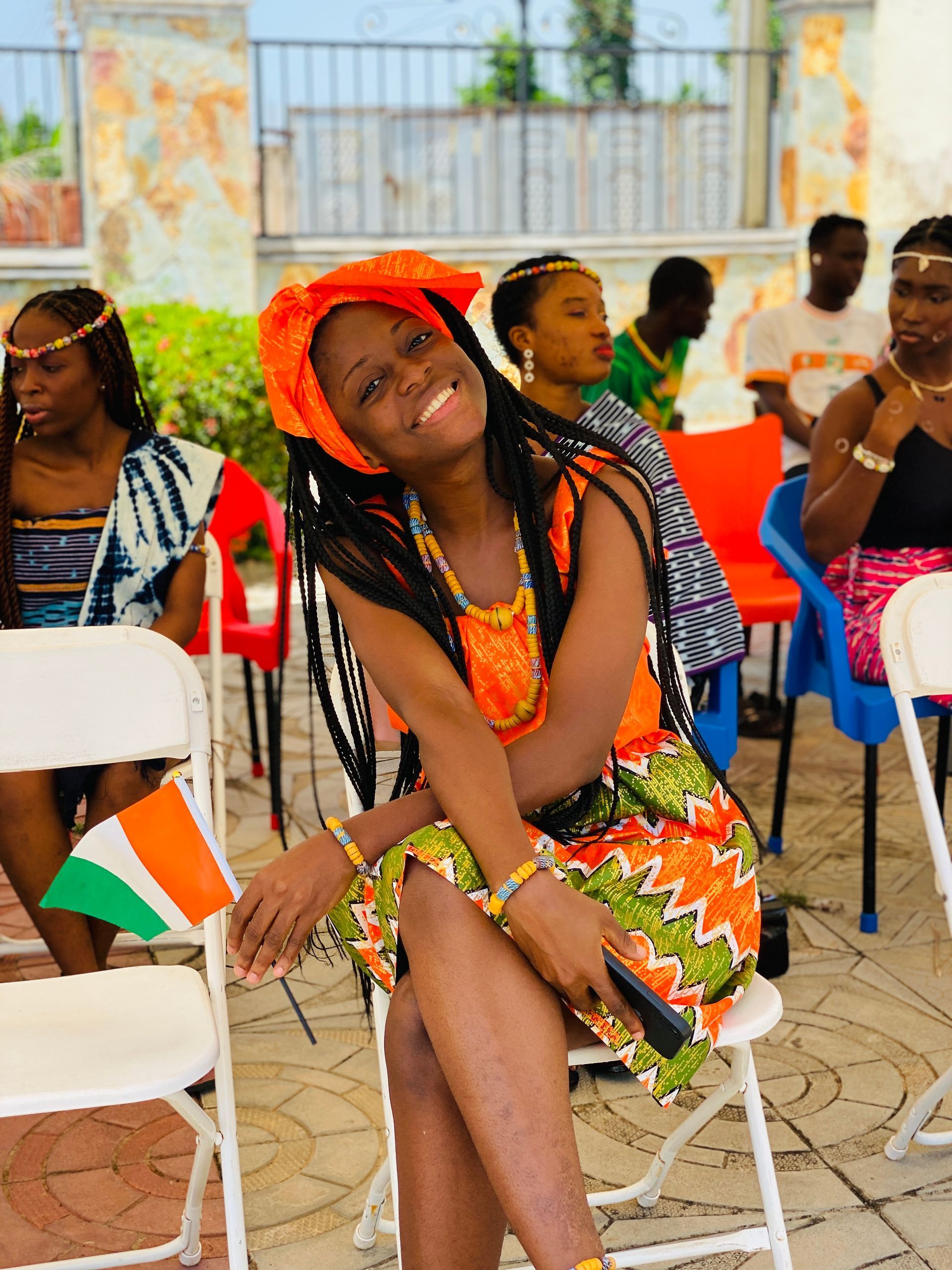 Une femme en robe traditionnelle orange et verte sourit en tenant un drapeau, assise à l'extérieur.