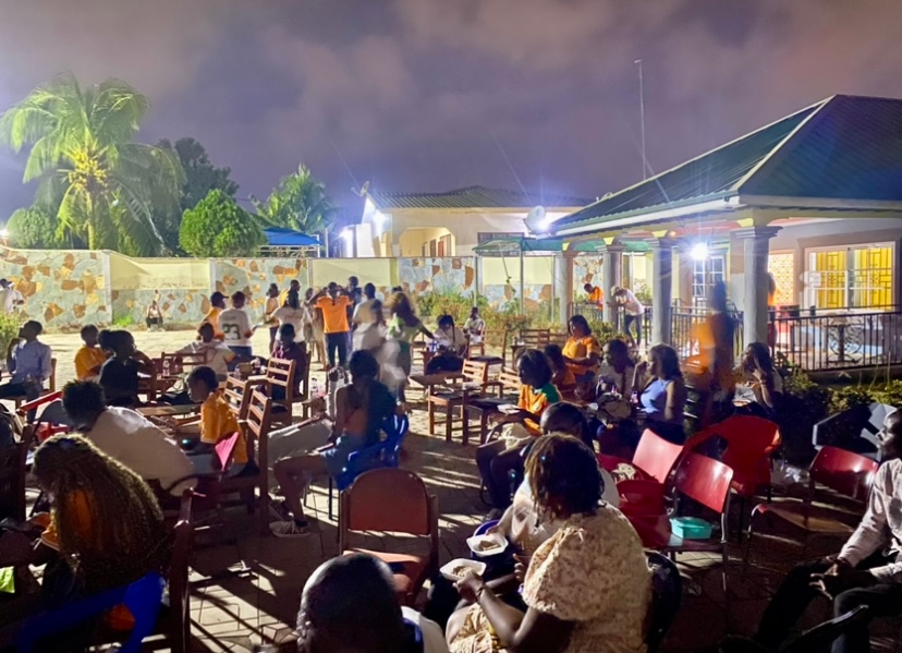 Rassemblement nocturne en plein air ; des gens discutent, certains dansent. Un bâtiment illuminé se profile à l’arrière-plan.