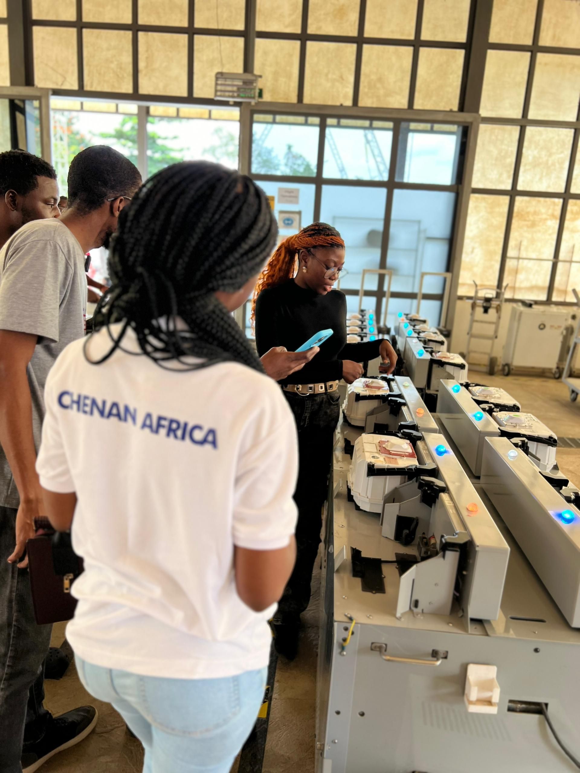 Des personnes examinent des machines dans une usine ; une femme porte un t-shirt « CHENAN AFRICA » et une autre personne utilise un téléphone.