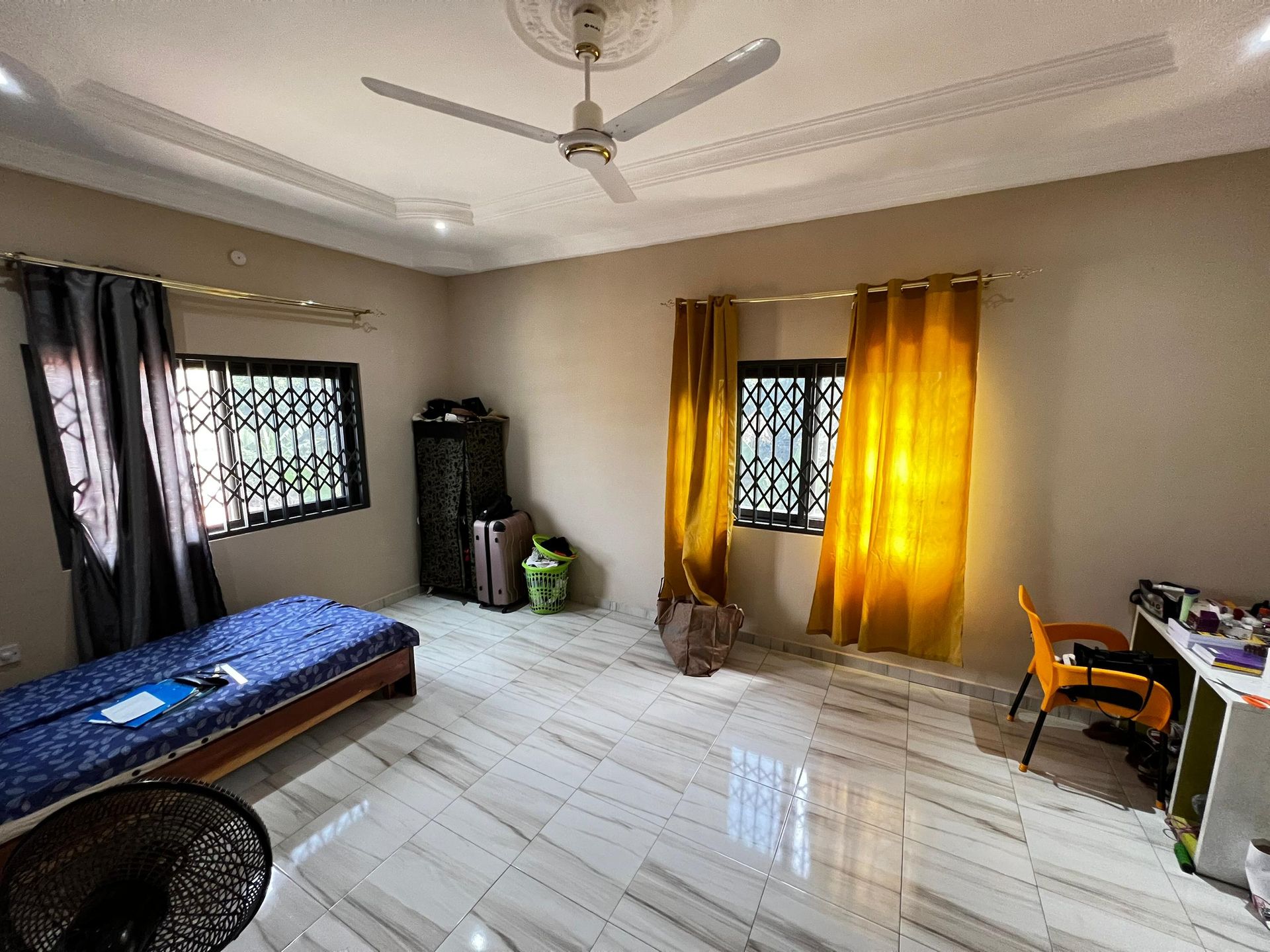 Chambre lumineuse avec lit, fenêtres, rideaux jaunes, ventilateur de plafond et bureau.