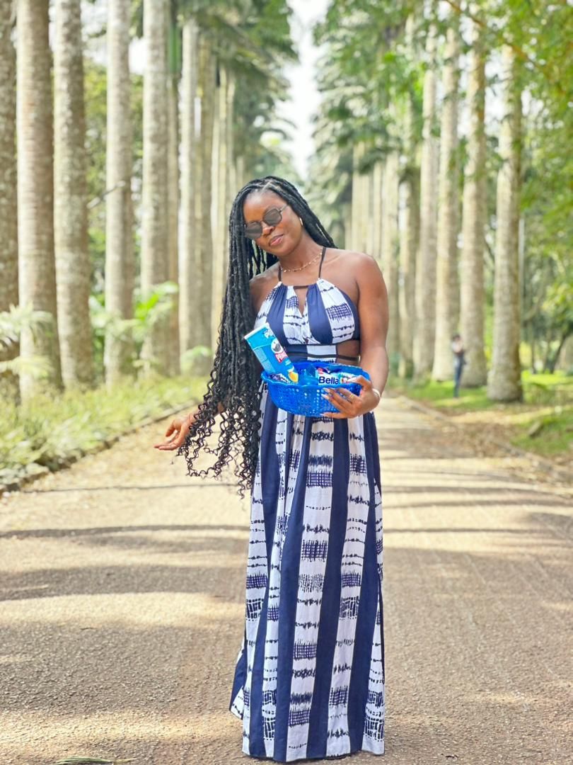 Une femme vêtue d'une robe à motifs bleus et blancs, tenant un cadeau, se tient sur un chemin bordé de grands arbres.