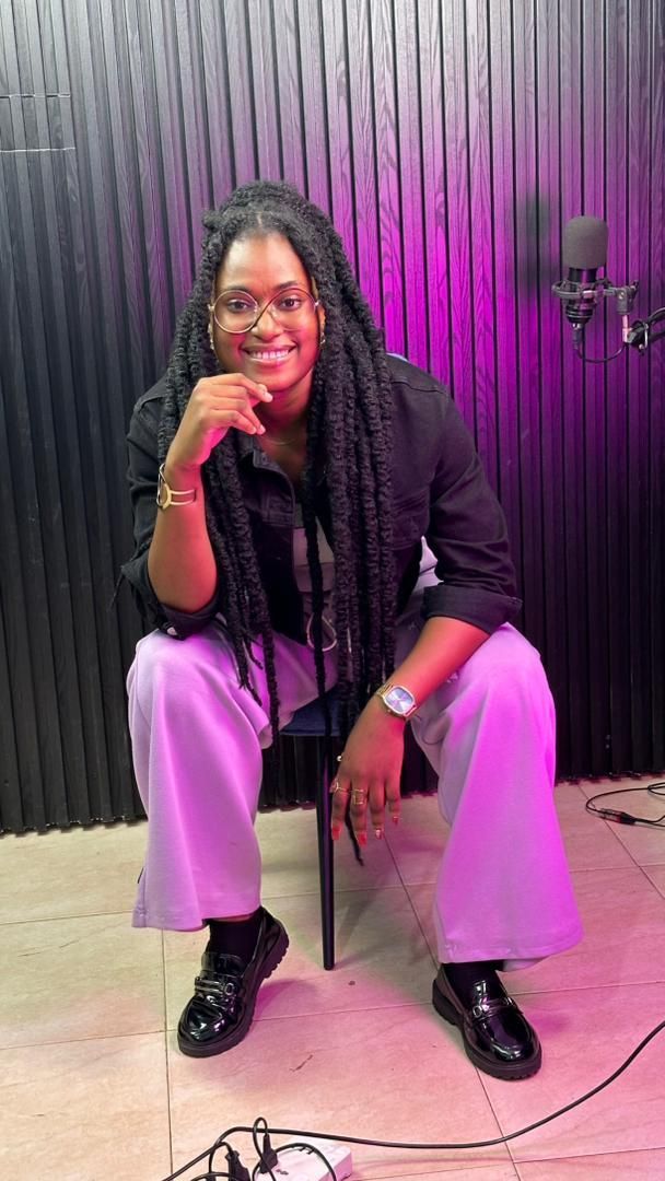 Une personne assise, souriante, dans un studio d'enregistrement. Elle porte des lunettes, des tresses, un haut noir et un pantalon lilas. Un microphone est présent.