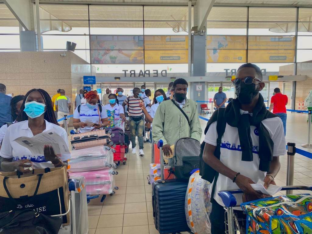 Des personnes avec des bagages dans un aéroport. Certaines portent des masques, font la queue et tiennent des documents.