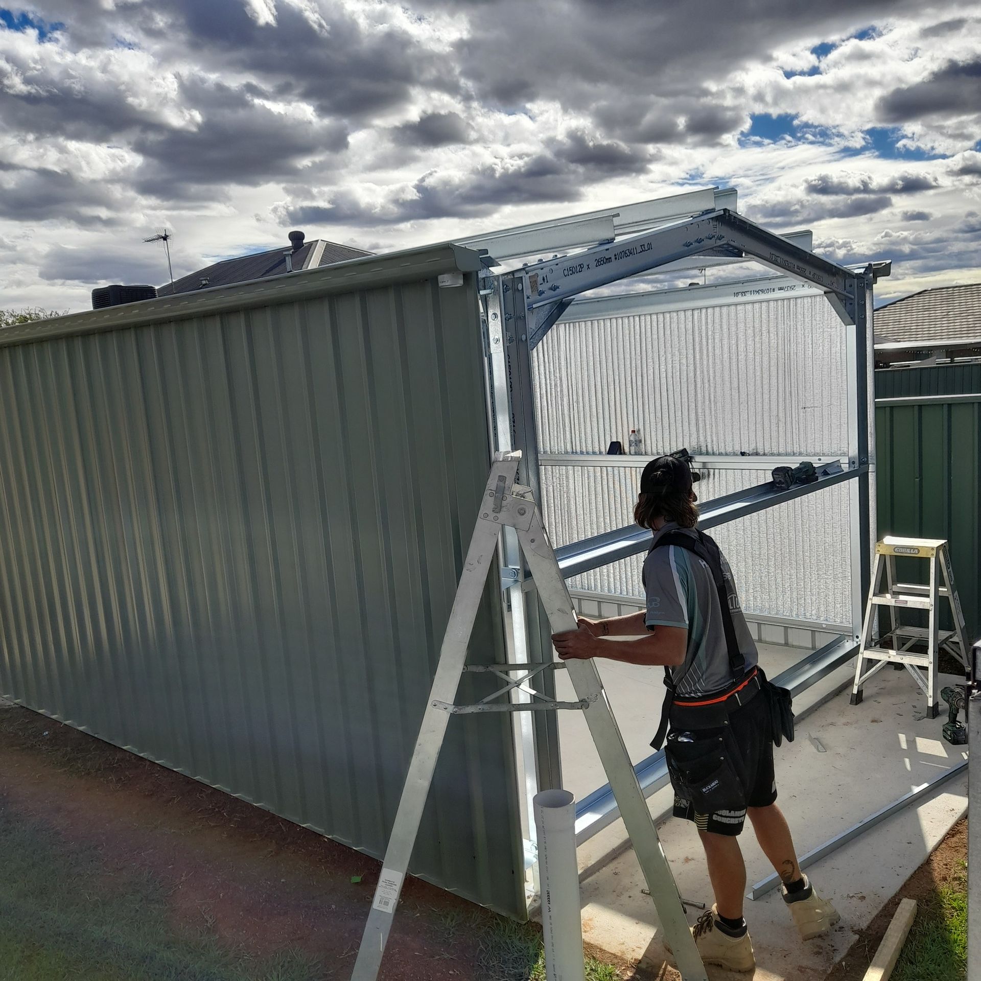 A worker on a ladder assembles a grey shed with corrugated metal siding under a cloudy sky — BJG Plumbing in Coolamon, NSW