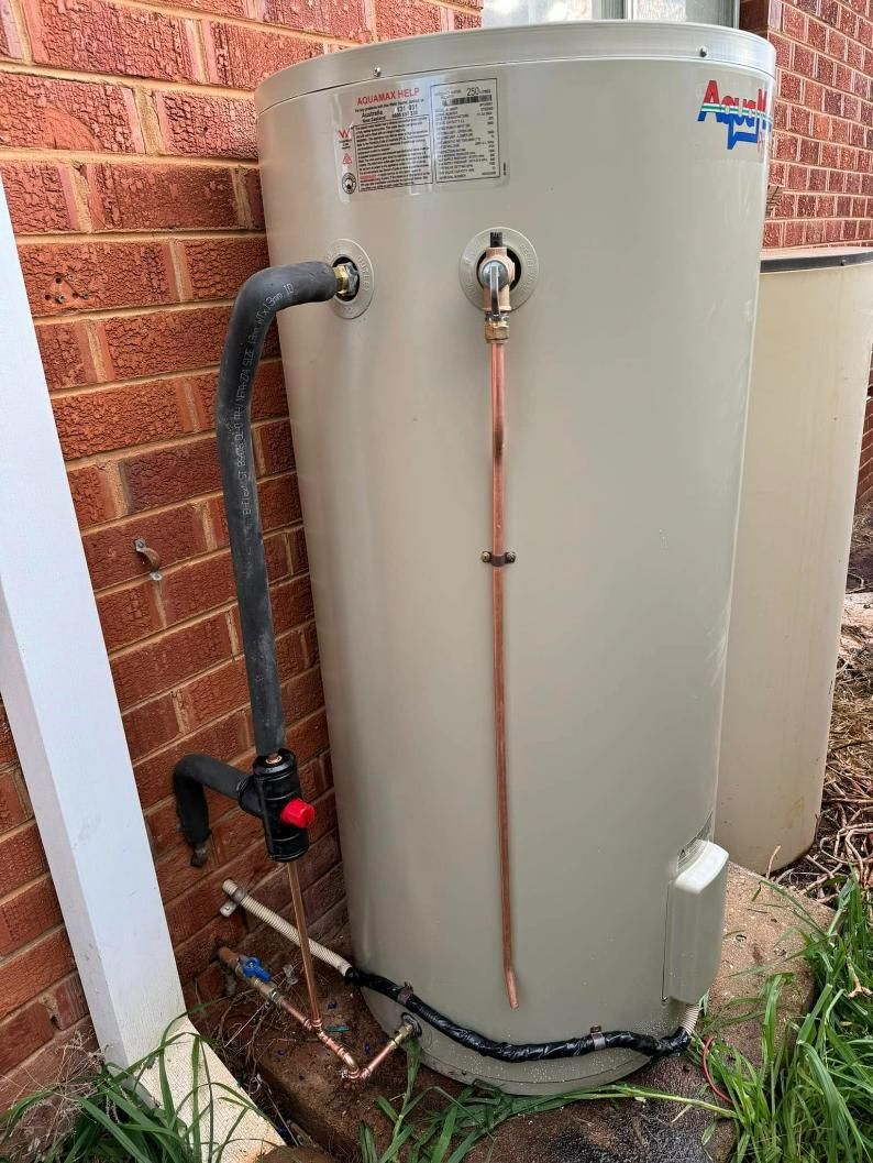 Tan Water Heater With Copper Pipes And Black Hose Beside Brick Wall And White Container — BJG Plumbing in Coolamon, NSW