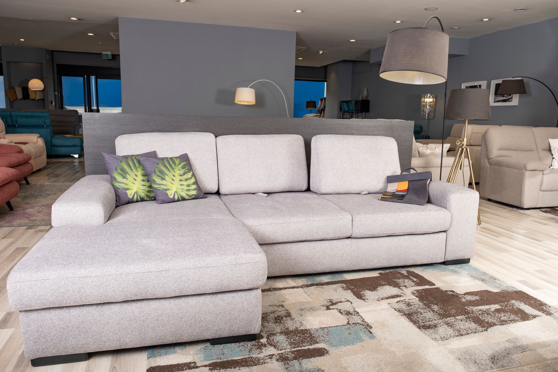 Divano componibile grigio con chaise longue in uno showroom, tappeto a fantasia e cuscini decorativi.