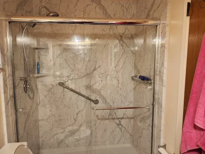 A walk-in shower with marble-patterned walls, a chrome grab bar, a handheld showerhead, and sliding glass doors.