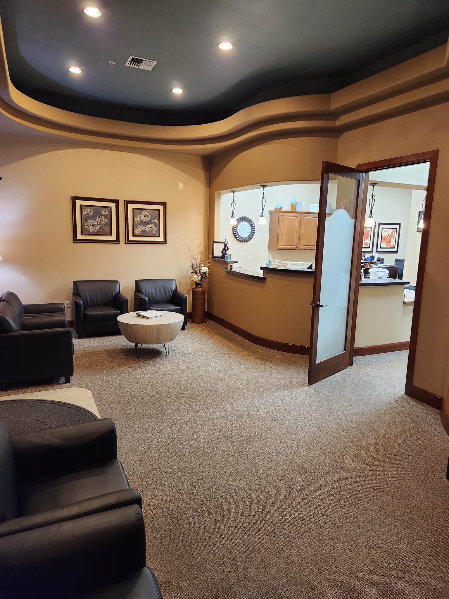 A waiting room with black armchairs, a round coffee table, carpeted flooring, and a receptionist desk through a doorway.
