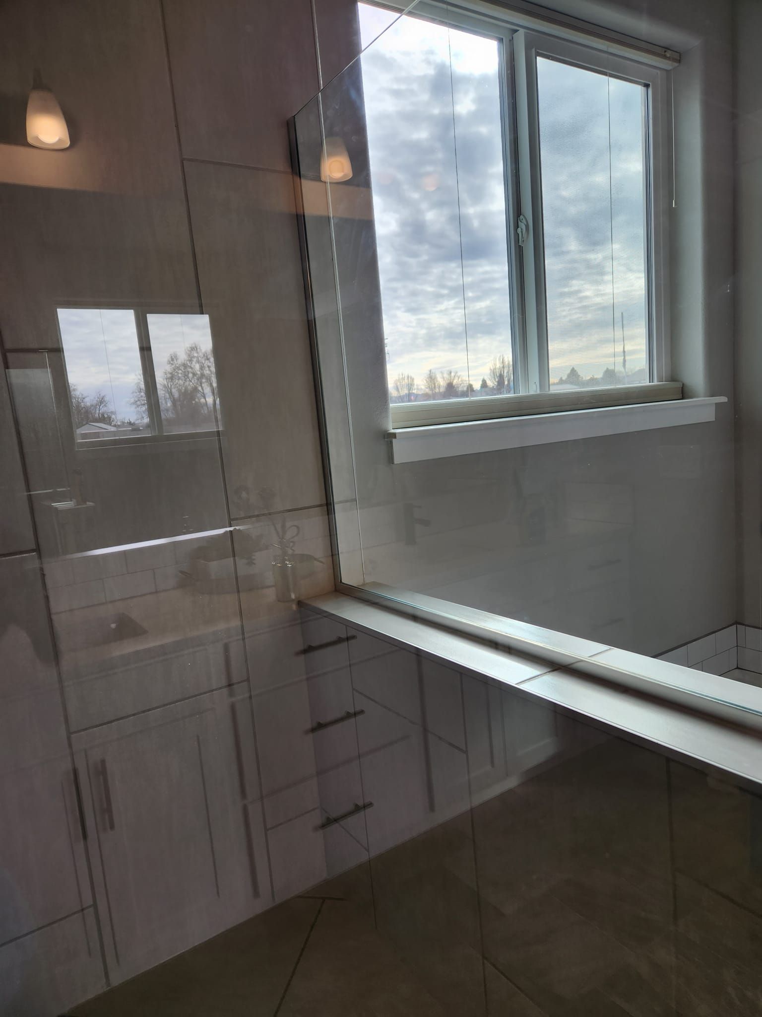 A window with a view of a cloudy sky above a tiled wall and a built-in narrow ledge in a modern bathroom.