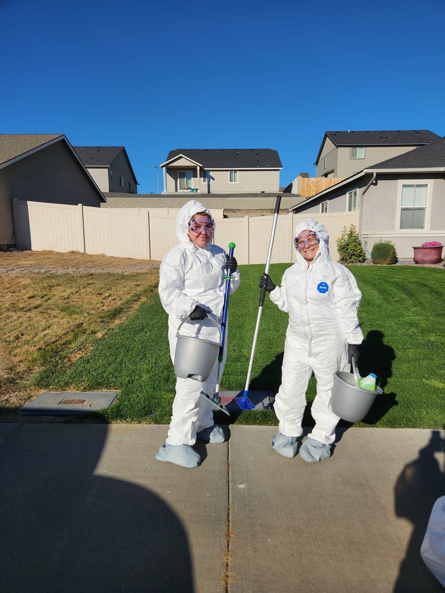 Two people in white protective suits and goggles stand on a sidewalk, holding paint rollers and buckets outside a home.