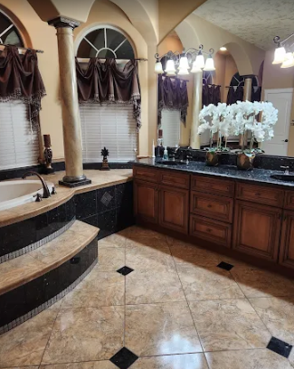 A luxurious bathroom with a corner tub, large wooden vanity, granite countertops, and stone tile floors with black accents.