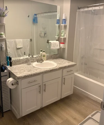 A clean bathroom with a white vanity, speckled countertop, sink, large mirror, and a walk-in shower with a white curtain.