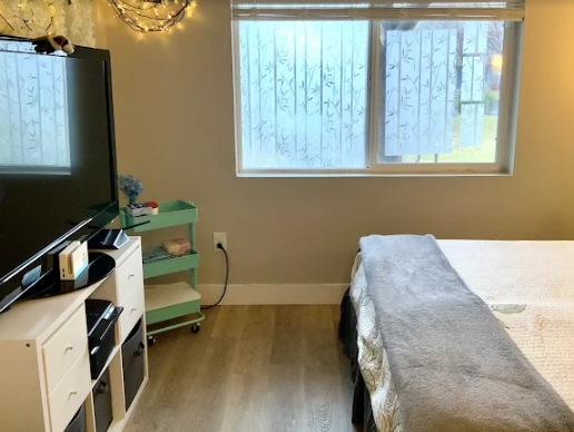 A bedroom corner with a television on a white storage unit, a teal rolling cart, and a bed with a grey blanket.