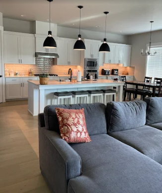 Open-concept kitchen with a gray sectional, kitchen island, and dining area, featuring white cabinets and pendant lights.
