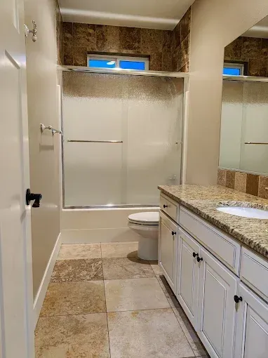 A beige bathroom with tile flooring, a granite-topped vanity, a toilet, and a glass-enclosed shower/tub combination.
