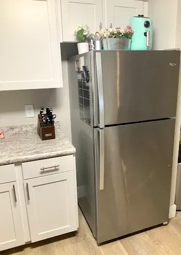 A stainless steel refrigerator sits next to white kitchen cabinets and a speckled countertop with a knife block.