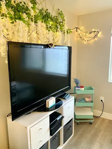 A large flat-screen TV on a white storage unit decorated with hanging floral vines and lights next to a teal rolling cart.