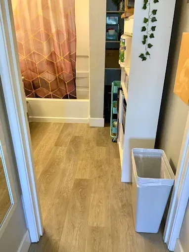 A view into a bathroom with wood-look flooring, a pink geometric shower curtain, shelving, and a white trash bin.