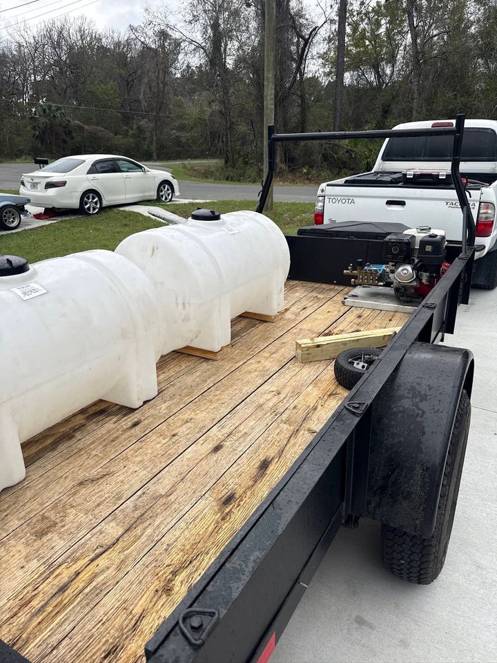 Black flatbed trailer loaded with white plastic tanks and equipment on a roadside in a wooded area