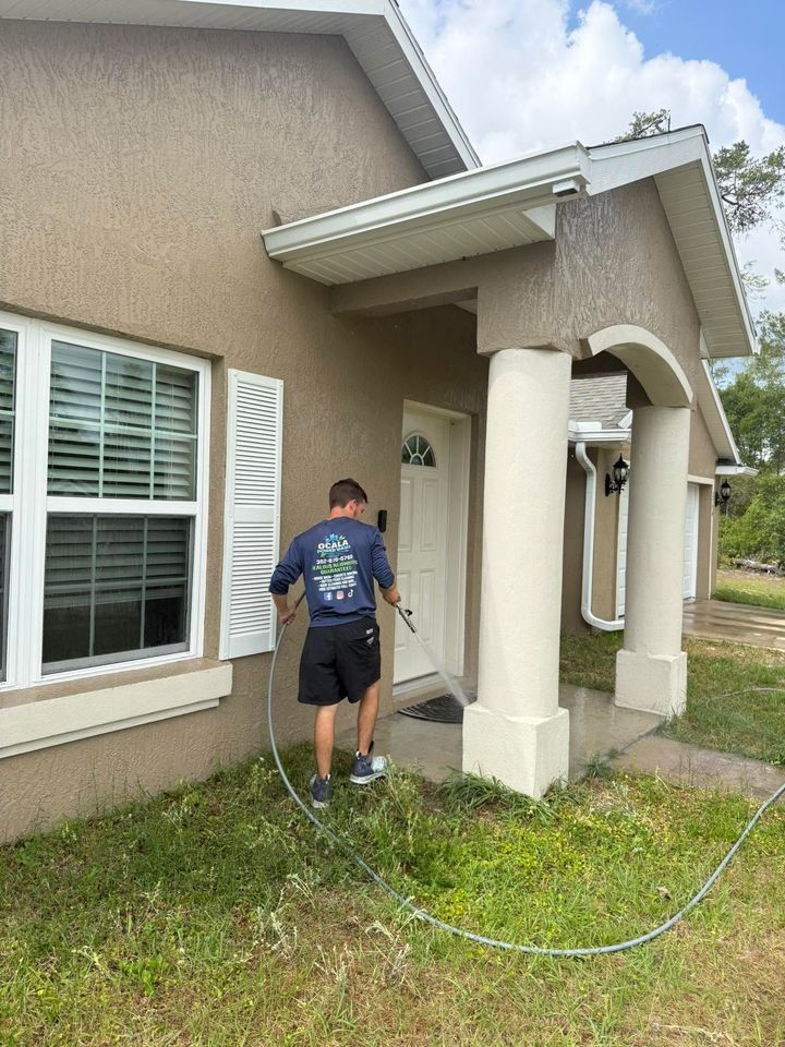 Worker pressure washing a stucco house exterior near a window and front porch columns