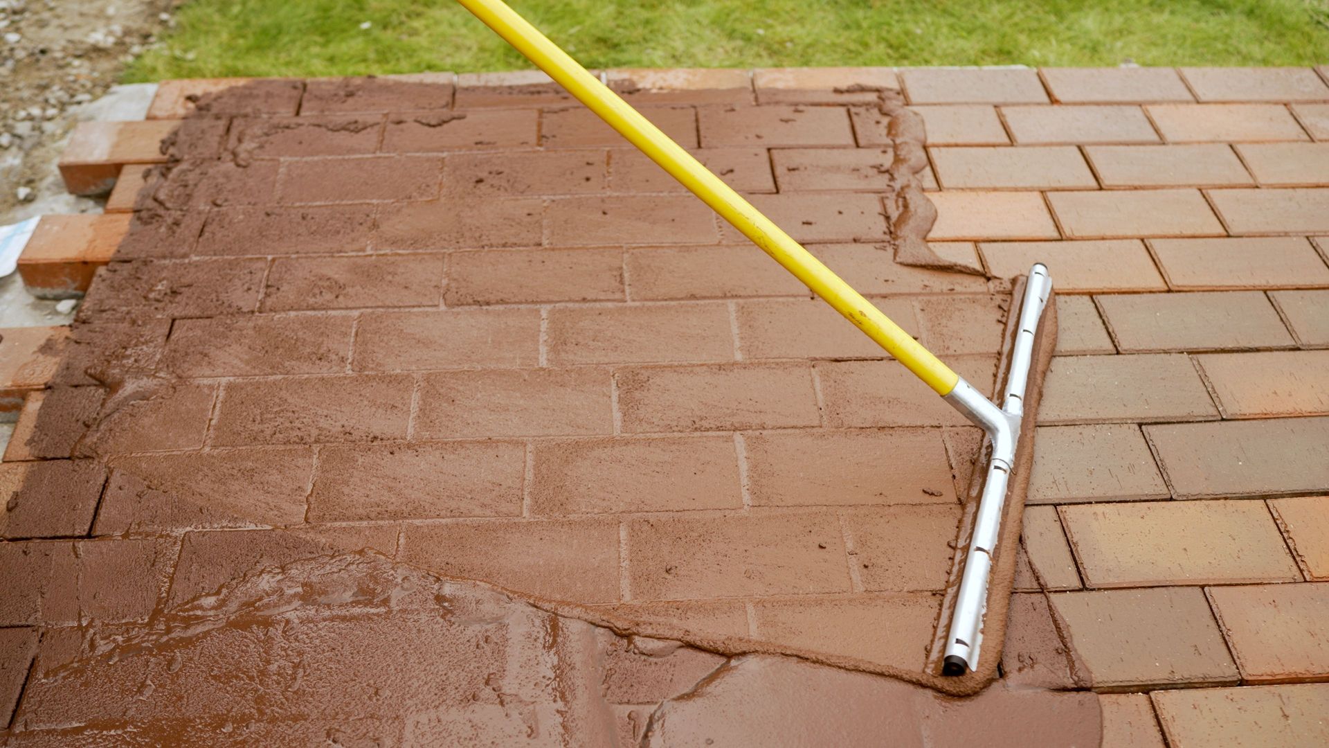 Yellow-handled floor squeegee smoothing wet brown concrete beside a brick walkway