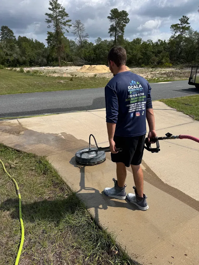 Person pressure-washing a concrete driveway beside a lawn and street on a sunny day