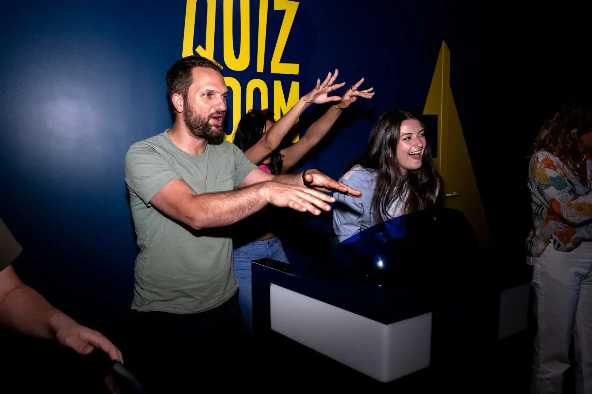 People in a quiz room, gesturing excitedly. Man in green shirt, woman with arms raised. Dark room, 